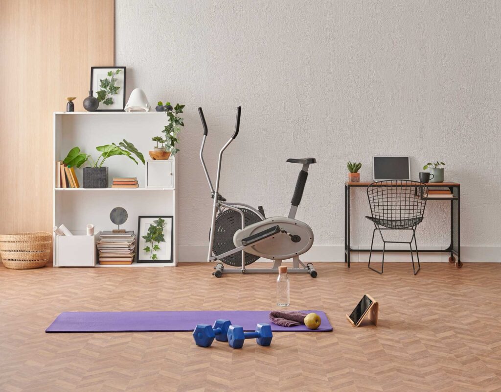 Home workout space with yoga mat, dumbbells, exercise bike, and desk setup in a modern living room
